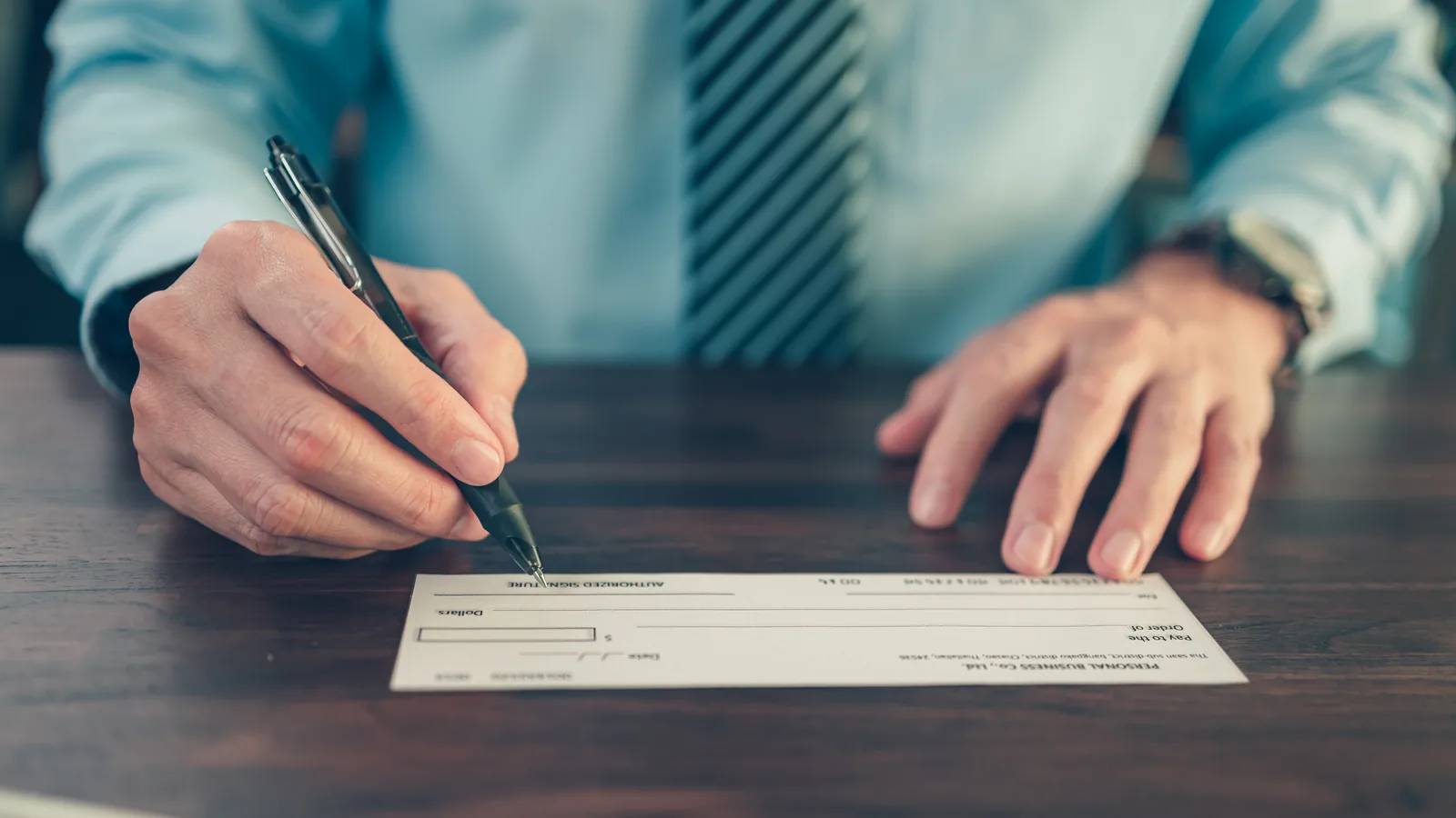 A businessman signing a blank bank cheque with a pen at a dark wooden desk.